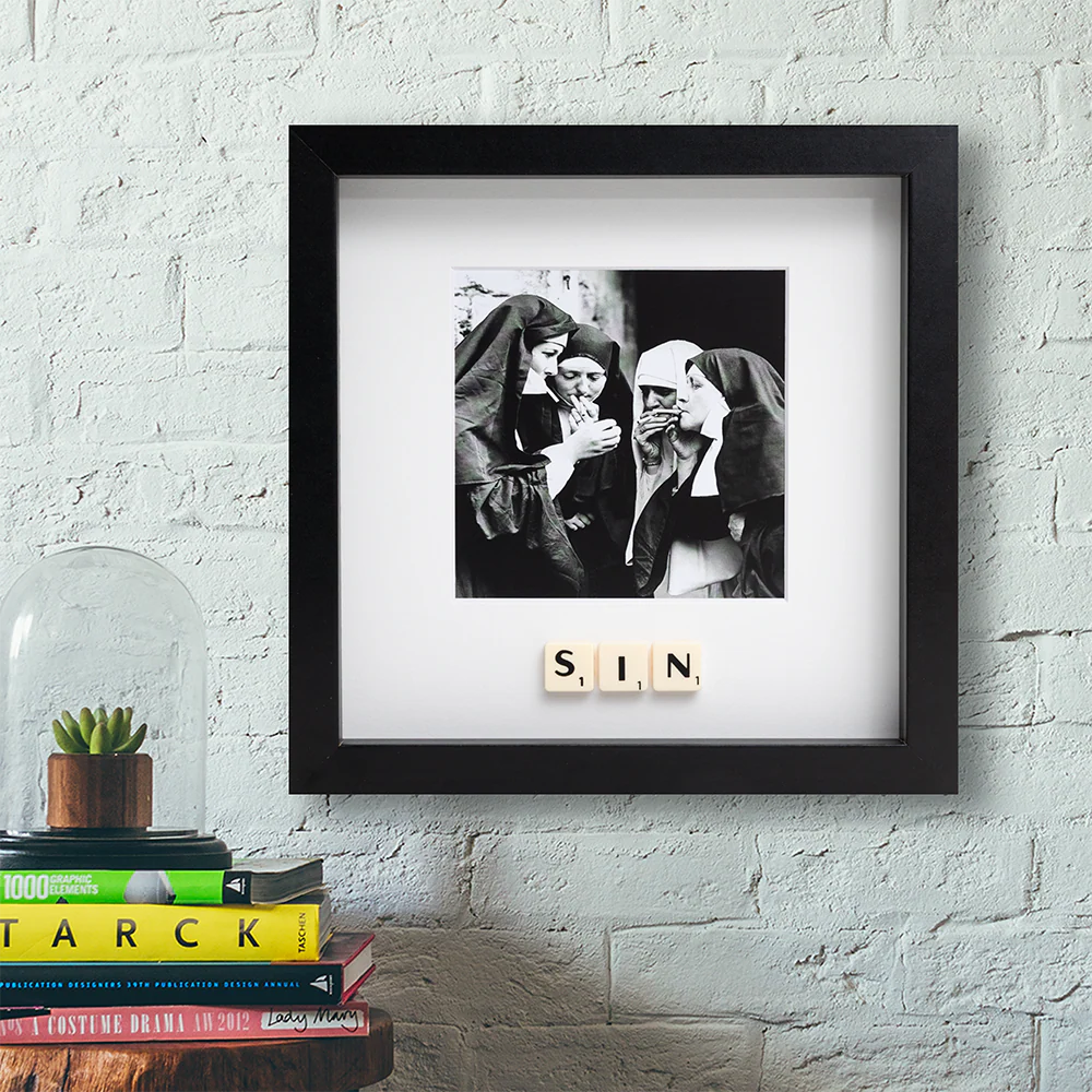 The SIN - Scrabble Print, framed in black and white, hangs on a white brick wall above a shelf with books and a glass cloche over a plant, capturing laughter and memory in its playful display. Gate21.ee