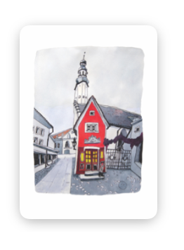 A small red building with glowing yellow windows stands in front of a larger white church with a tall spire. “Fuck - Tea-towels” hang in the window, adding a cosy touch to the quiet street scene beneath the overcast skies. Gate21.ee