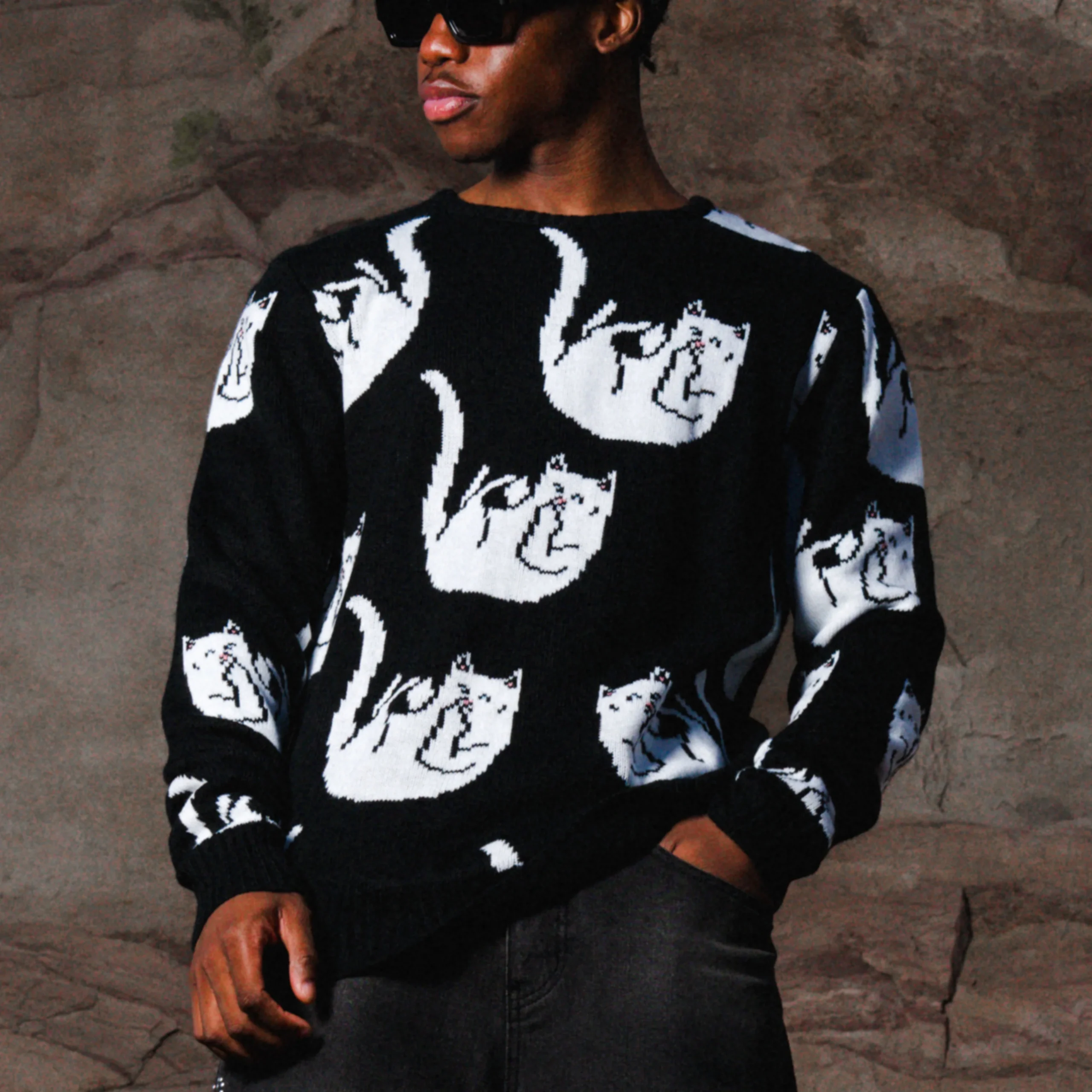 A person wearing sunglasses and the black Falling for Nermal jumper by Ripndip, featuring a repeated white cat design, stands with hands in pockets against a brown textured background. Gate21.ee
