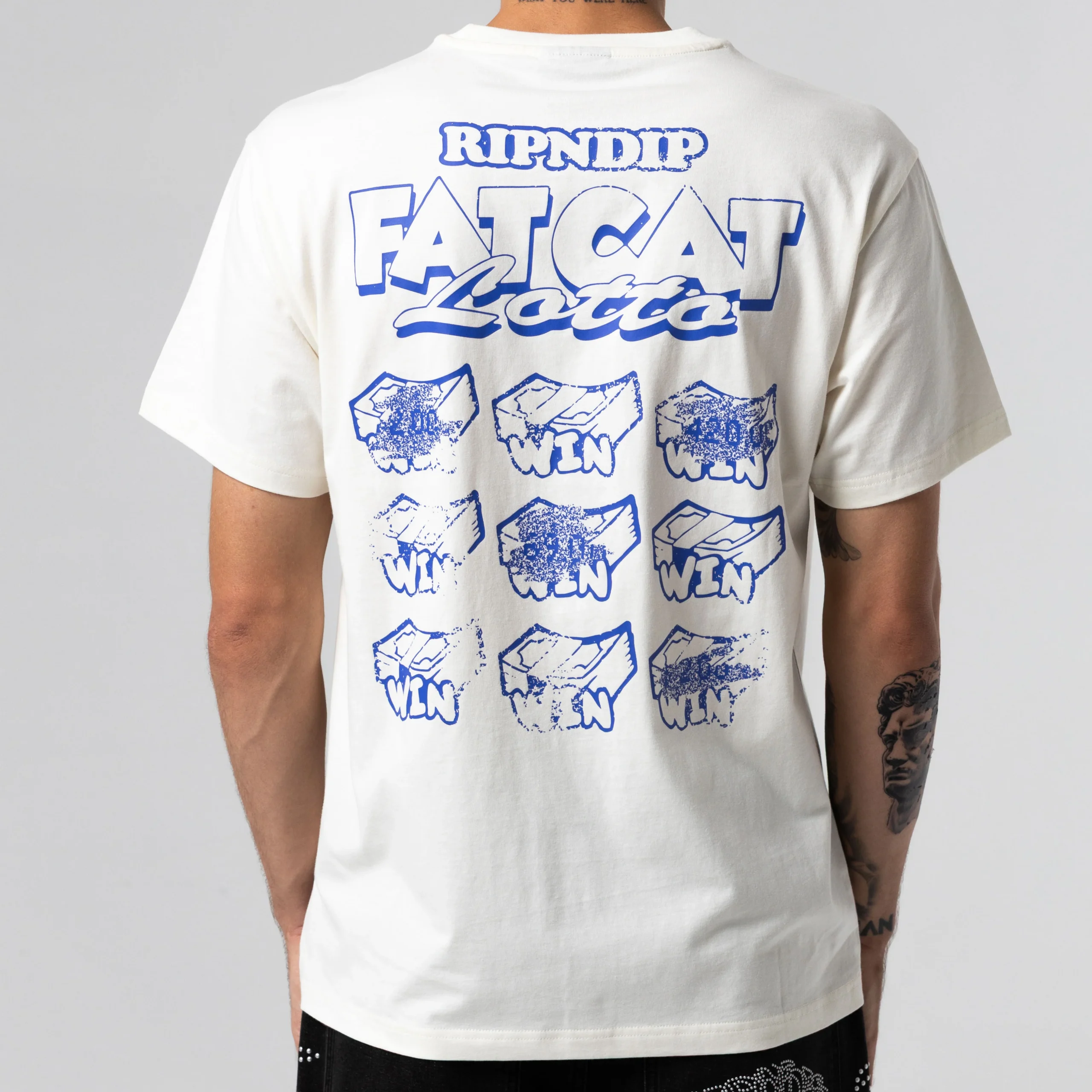 Wearing the Fat Cat Tee - Bone, which features blue graphics and “RIPNDIP FATCAT Lotto” above a scratch card grid with all “WIN,” a person stands with their back to the camera. Gate21.ee