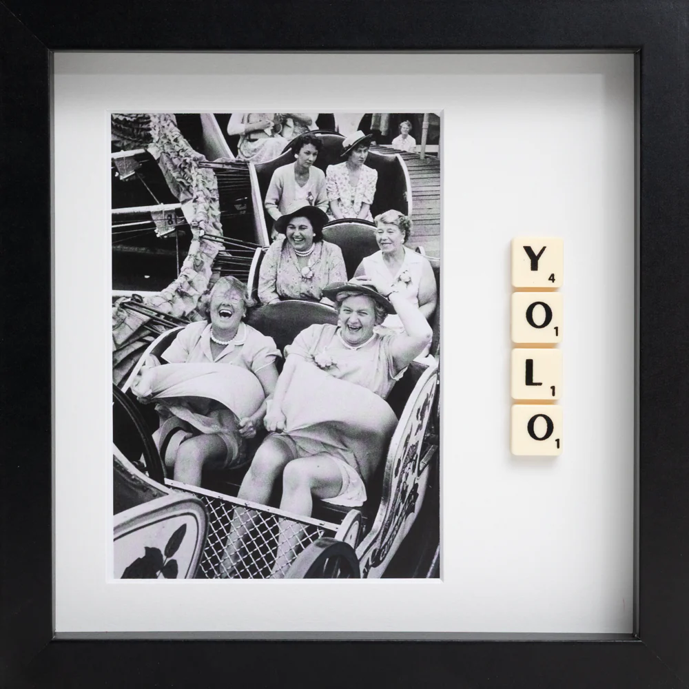 The YOLO - Scrabble Print features a framed black-and-white photo of women laughing on a rollercoaster, with Scrabble tiles spelling "YOLO"—a perfect piece to spark memories and celebrate moments that matter. Gate21.ee