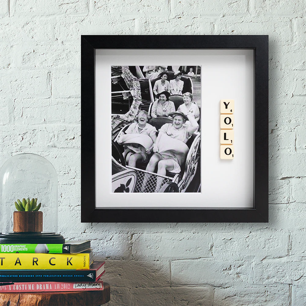 The YOLO - Scrabble Print is a framed black and white photo of people on a funfair ride, with “YOLO” spelt in Scrabble tiles. It hangs on a white brick wall above books and a small plant, adding inspiration to your space. Gate21.ee