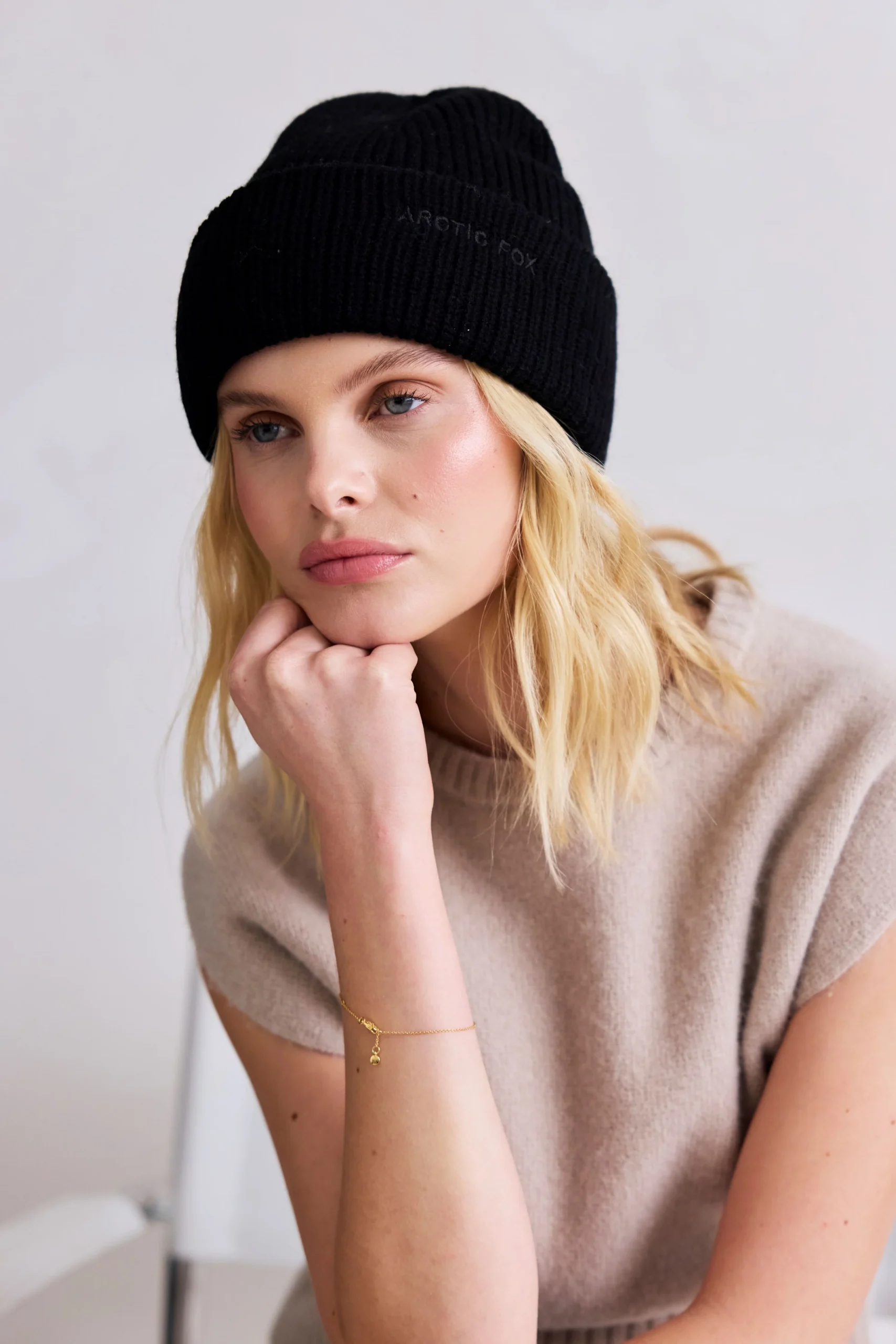 A woman with blonde hair wears the Arctic Black – Merino Beanie, a beige sleeveless top, and a gold bracelet. She rests her chin on her hand and gazes forward with a neutral expression against a plain, light background. Gate21.ee