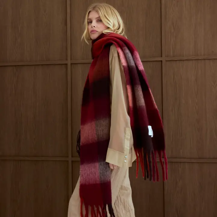 A person with fair hair, dressed in beige and wearing the Reykjavik Scarf - Burgundy Check, stands sideways in front of a wooden panelled wall. Gate21.ee