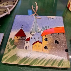 The Helina Tilk V&auml;ike alus is a ceramic tile with a colourful, stylised illustration of Tallinn&rsquo;s old town&mdash;towers, rooftops, and church spire&mdash;with a hanging string through a small hole, displayed on a green surface. Gate21.ee