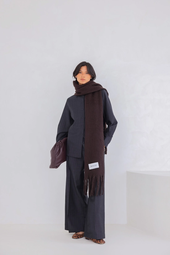 A person stands against a plain white background wearing a dark button-up shirt, wide-leg trousers, leopard print shoes, and the Reykjavik Scarf in Ground Coffee, holding a dark-coloured bag under one arm. Gate21.ee