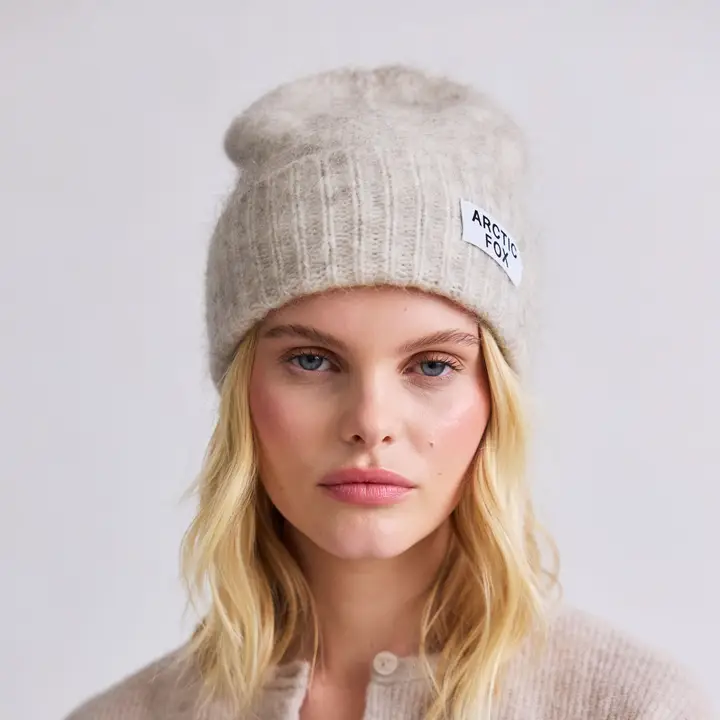 A person with long blonde hair wears the Mohair Beanie in Alpine Ecru and a matching button-up cardigan, standing against a plain light background. Gate21.ee