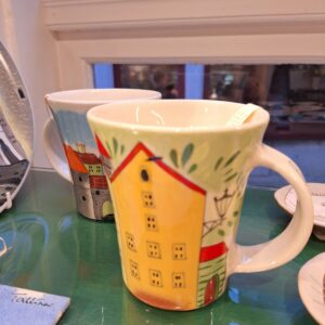 The Helina Tilk Tass ceramic mug, featuring a colourful yellow building illustration, sits on a green glass surface by a window. Behind it, another mug from the collection with a different building design is partially visible. Gate21.ee