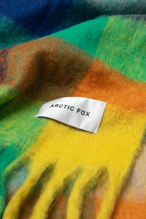 A close-up of the Reykjavik Scarf - Nordic Skies shows its fuzzy, multicoloured fabric in green, yellow, and blue hues. A white label sewn onto it features ARCTIC FOX in bold black capitals. Gate21.ee