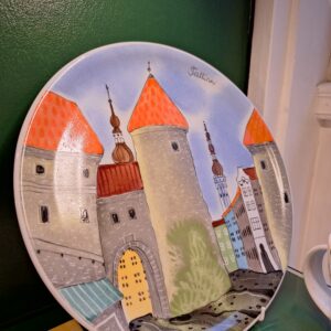 The Helina Tilk Suur taldrik is a decorative plate showcasing an illustrated cityscape with stone towers, red-orange roofs, colourful buildings, and the word "Tallinn" near the top edge. Displayed on a shelf. Gate21.ee