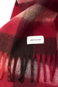 A close-up of The Reykjavik Scarf - Burgundy Check, showing its red and black check pattern, white label, and fringed edge at the bottom. Gate21.ee