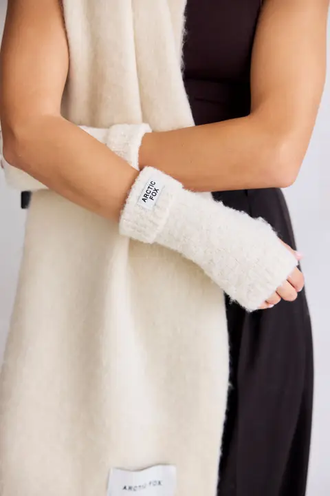 A person in a sleeveless black top models cream-coloured Reykjavik Hand Warmers - Pebble Beige by Arctic Fox and a matching scarf, both with small Arctic Fox labels, arms crossed in front of their body. Gate21.ee