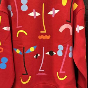 A Red - Pusa hoodie by RCR Khomenko hangs against a black background, showcasing an abstract design with colourful geometric shapes and lines in blue, yellow, pink, black, and white that resemble stylised faces. Gate21.ee