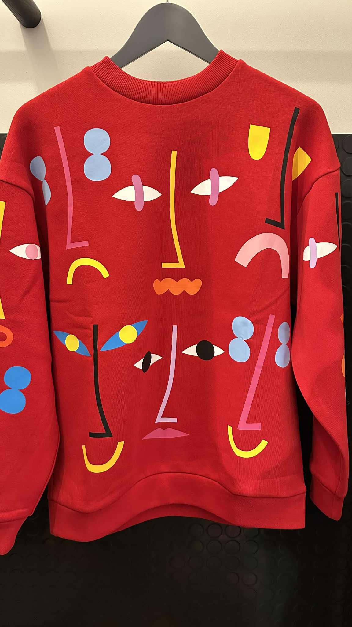 A Red - Pusa hoodie by RCR Khomenko hangs against a black background, showcasing an abstract design with colourful geometric shapes and lines in blue, yellow, pink, black, and white that resemble stylised faces. Gate21.ee