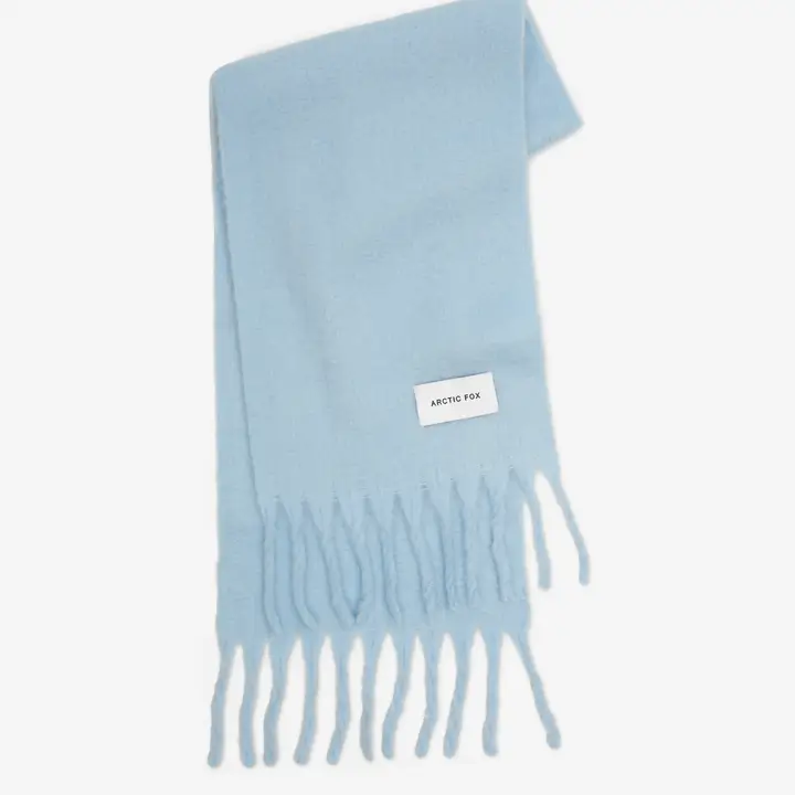 The Stockholm Scarf - Glacier Blue is a light blue scarf with fringed ends and a white rectangular tag reading ARCTIC FOX STOCKHOLM, shown laid flat on a white background. Gate21.ee