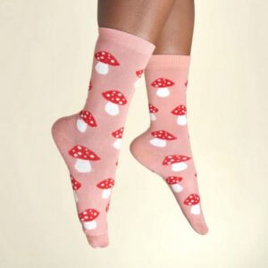 A pair of feet wearing Mushroom - Cotton Socks by Coucou Suzette, featuring a red and white mushroom pattern on pink cotton, set against a plain light beige background. Gate21.ee