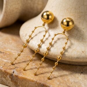 The Vivienne - BØLD Earrings feature gold-tone drops with round studs and two arched, beaded chains on each, shown on a textured stone surface. Gate21.ee