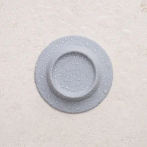 The Soapi - Grey, a round light grey silicone object with a raised centre and water droplets on its surface, rests on a textured light beige background. Gate21.ee