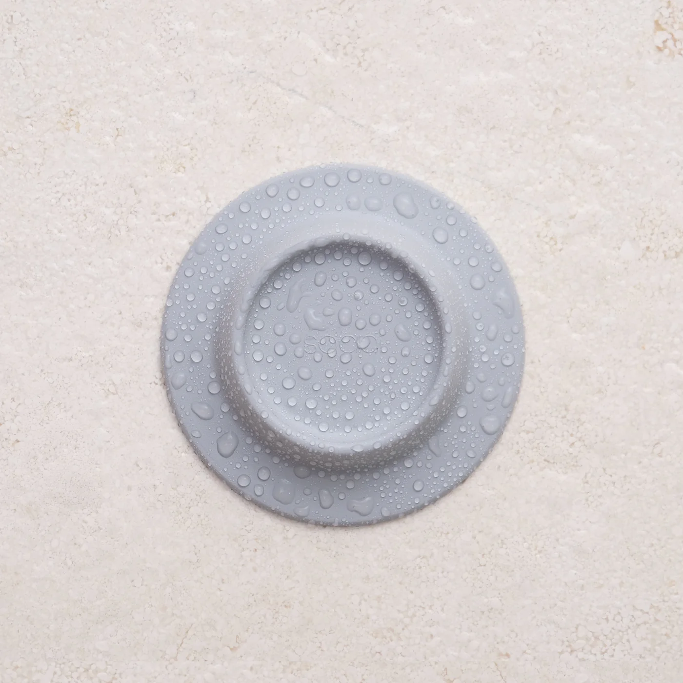 The Soapi - Grey, a round light grey silicone object with a raised centre and water droplets on its surface, rests on a textured light beige background. Gate21.ee