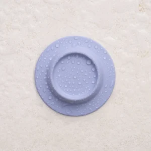 The Soapi - Lavender is a round, flat, light purple soap with a raised edge and water droplets on its surface, set against a textured beige background. Gate21.ee
