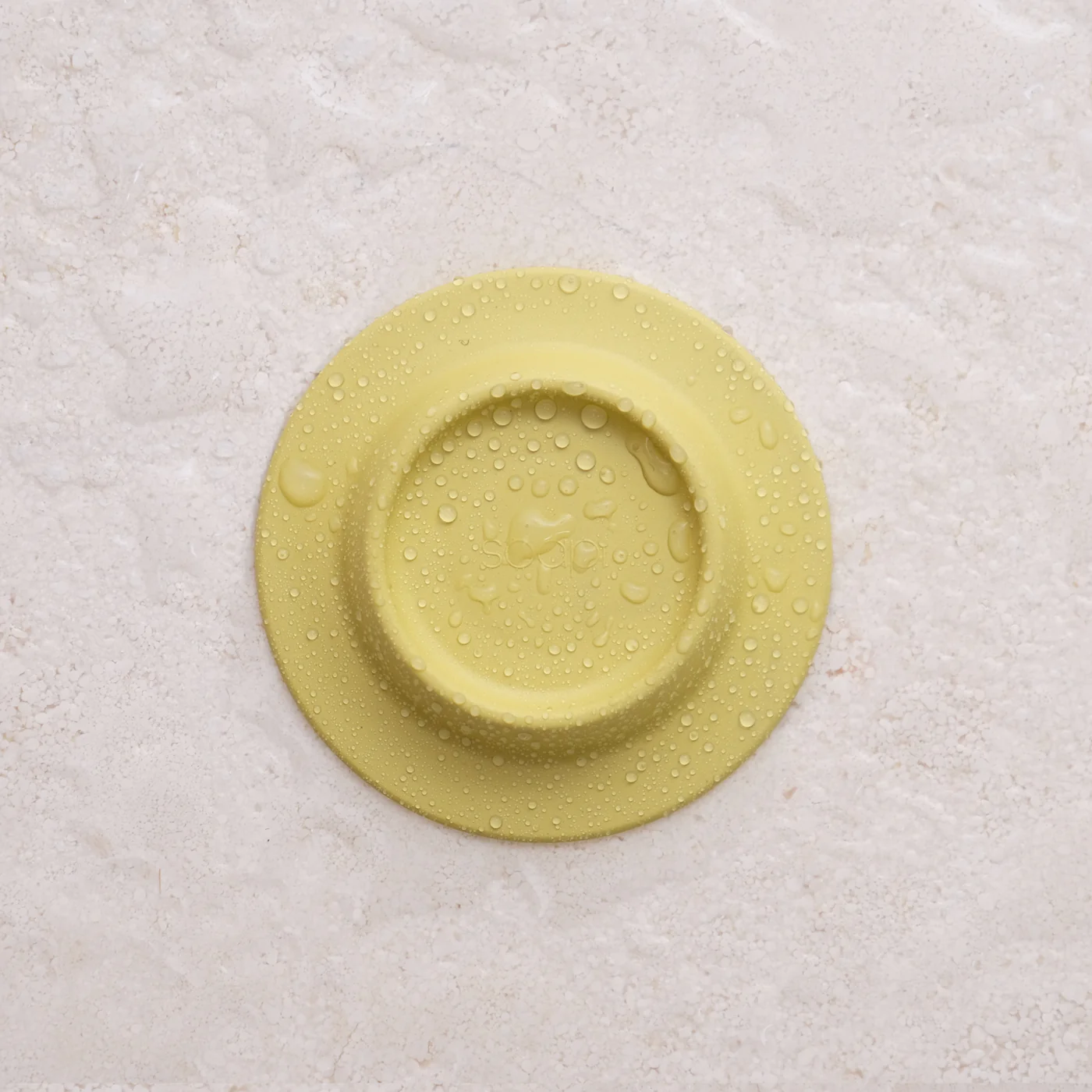 The Soapi - Yellow silicone object, featuring a round design and water droplets on its surface, is shown on a light textured background. Gate21.ee