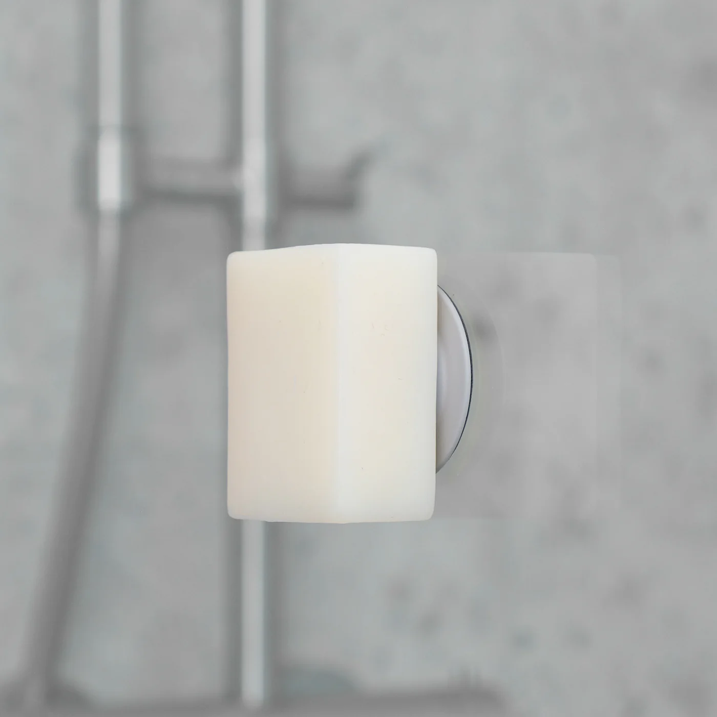 A rectangular grey Soapi bar of soap is mounted on a circular holder attached to a bathroom wall, with a blurred shower fitting in the background. Gate21.ee
