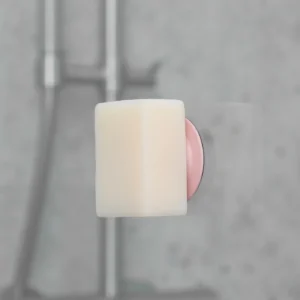The Soapi - Pink, a wall-mounted pink and silver soap holder, holds a rectangular white bar of soap in a bathroom with a blurred grey tiled background. Gate21.ee