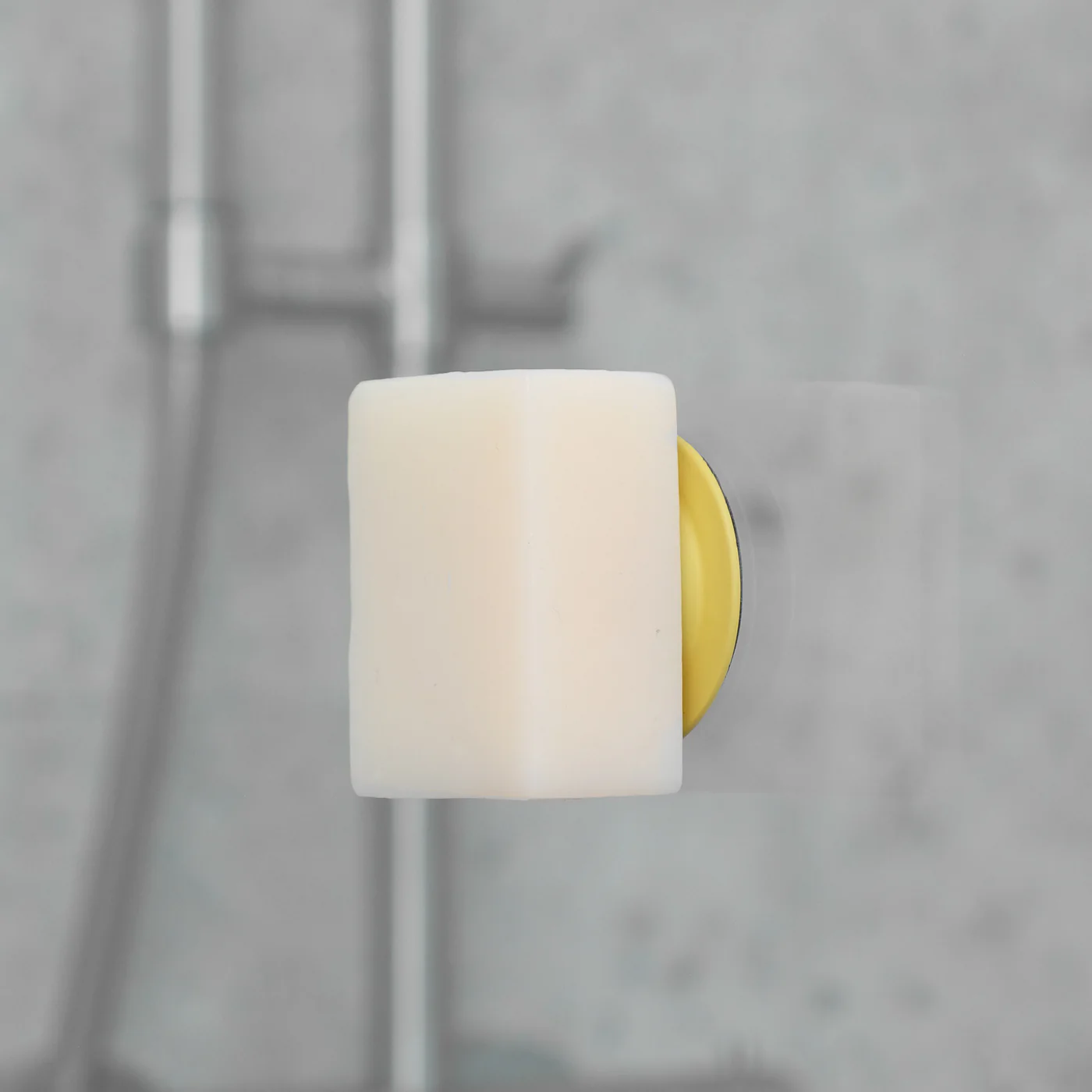 A rectangular white Soapi - Yellow bar sits in a yellow and clear wall-mounted holder in a bathroom, with a blurred shower fitting visible in the background. Gate21.ee