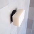 A rectangular white soap bar is held by a round, black Soapi - Black wall-mounted holder on a tiled bathroom wall, with a shower curtain partially visible to the right. Gate21.ee