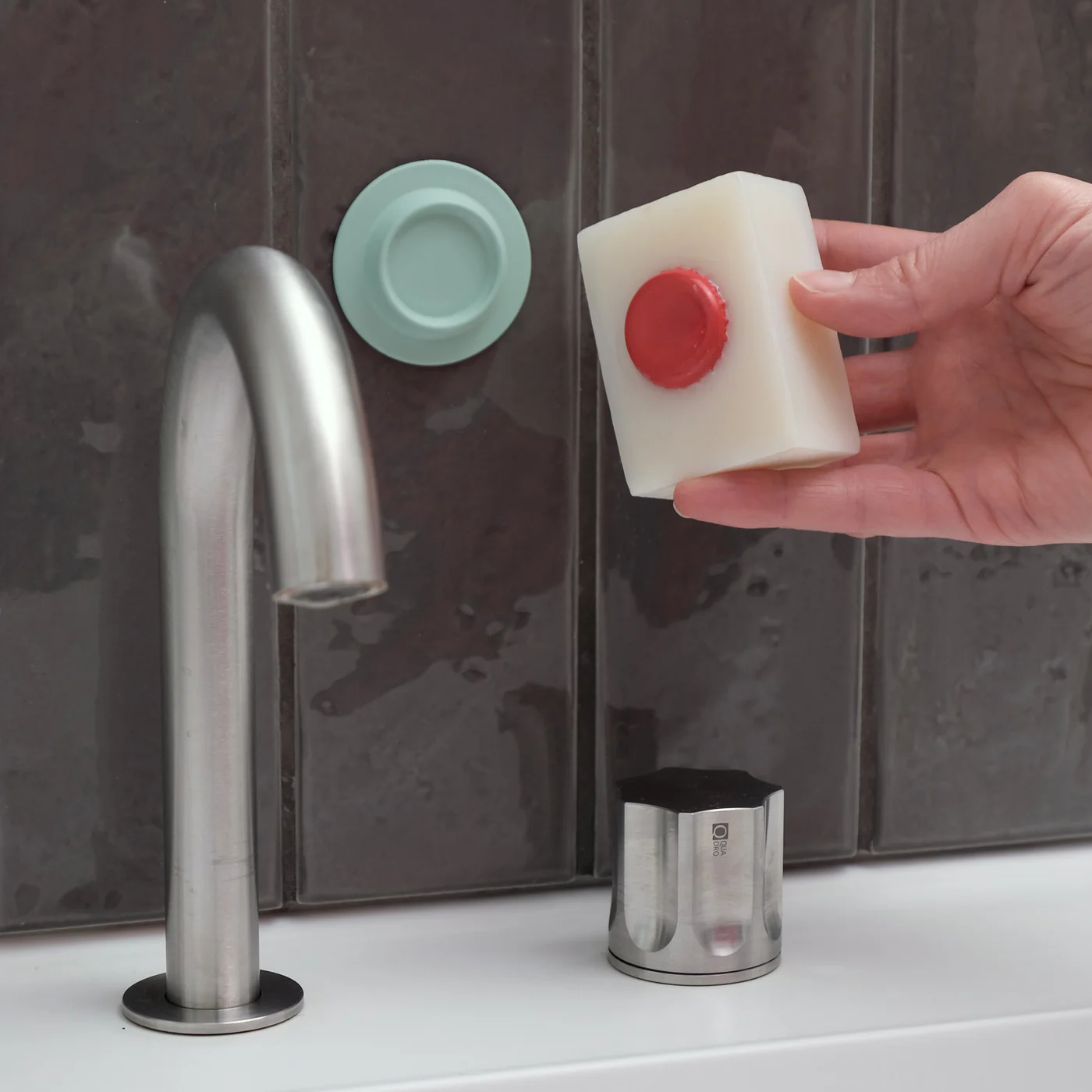 A hand holds a rectangular Soapi - Off White soap with a red button near a mint green wall holder above a silver tap on a white basin. Gate21.ee