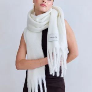 A person with fair hair wears the Stockholm Scarf - Polar White from Arctic Fox over a sleeveless black outfit. The thick, fringed scarf features an ARCTIC FOX patch. The person looks slightly to the side against a plain background. Gate21.ee