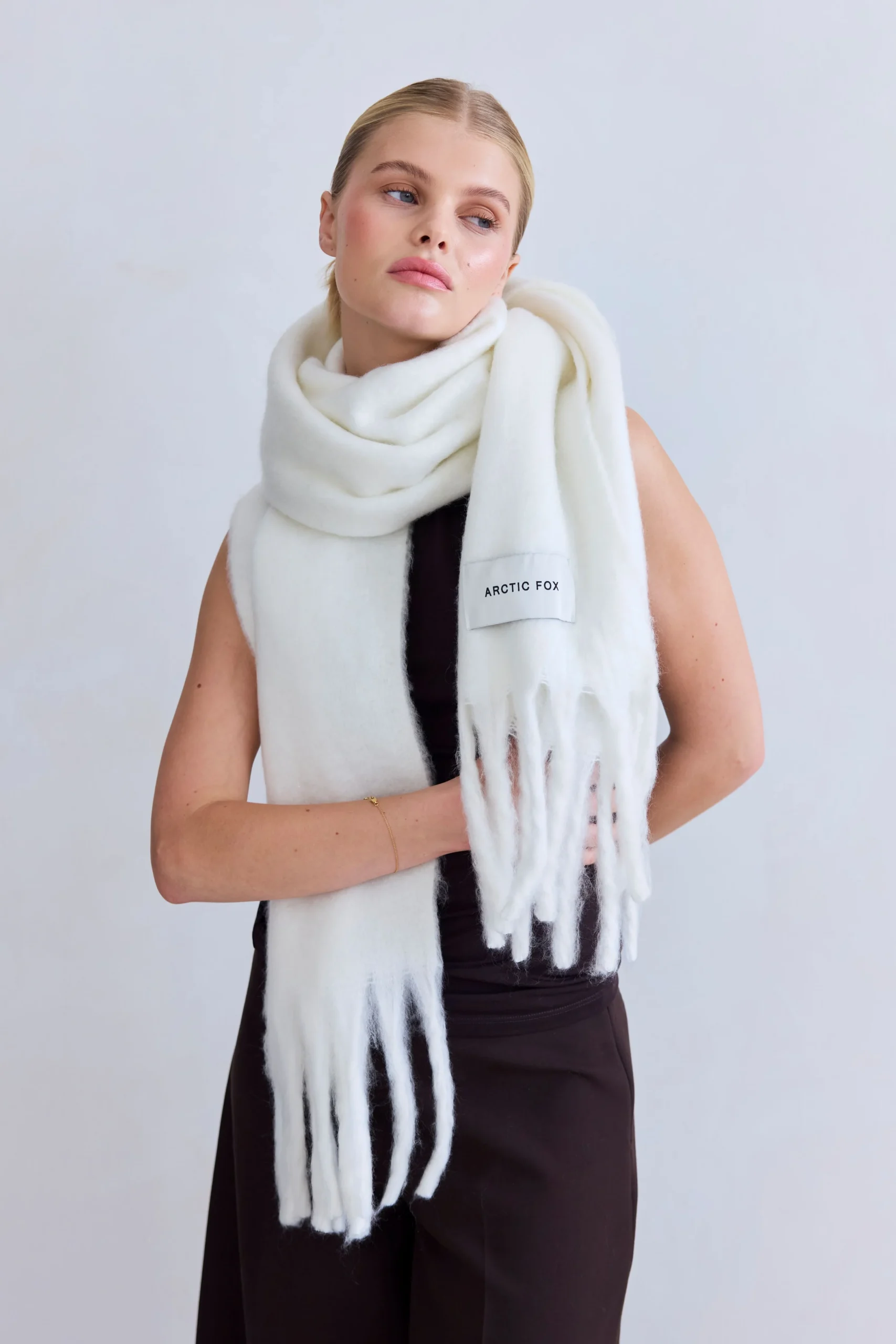 A person with fair hair wears the Stockholm Scarf - Polar White from Arctic Fox over a sleeveless black outfit. The thick, fringed scarf features an ARCTIC FOX patch. The person looks slightly to the side against a plain background. Gate21.ee