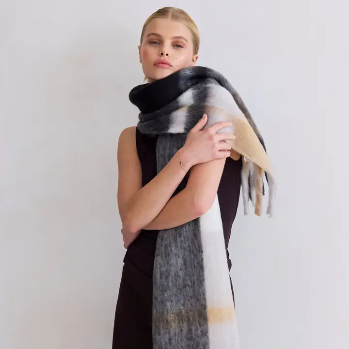 A woman in a sleeveless black dress stands with arms crossed, wearing the Stockholm Scarf - the Arctic, a long and thick scarf featuring black, grey, white, and beige stripes against a plain light background. Gate21.ee