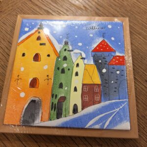 The Helina Tilk Suur alus ceramic tile displays vibrant, tall, narrow buildings with arched doors and windows on a snowy backdrop. Wrapped in plastic, it rests on a wooden surface. Gate21.ee