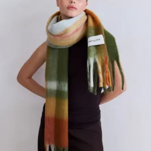 Wearing a sleeveless black top and long dark skirt, a model stands against a light background showcasing the Stockholm Scarf - Mossy Spring, featuring green, orange, brown, and white tones with an Arctic Fox Stockholm tag. Gate21.ee