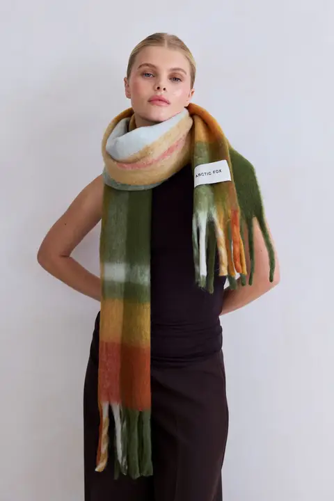 Wearing a sleeveless black top and long dark skirt, a model stands against a light background showcasing the Stockholm Scarf - Mossy Spring, featuring green, orange, brown, and white tones with an Arctic Fox Stockholm tag. Gate21.ee