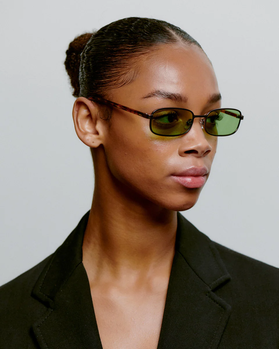 A person with slicked-back hair in a bun wears NOAH - Gun Metal Havana sunglasses and a black blazer. The background is plain and light-coloured, and they are looking slightly to the left. Gate21.ee