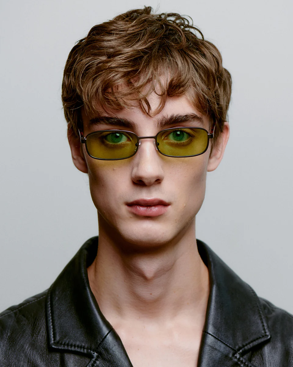 Young person with short, tousled brown hair wears NOAH - Gun Metal Havana sunglasses from A. Kjærbede and a black leather jacket, looking directly at the camera against a plain light background. Gate21.ee