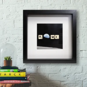 The HOME - Scrabble Print features a framed photo of Earth from the Moon, with Scrabble tiles spelling "HOME," displayed on a white brick wall above a stack of books and a small plant in a glass dome. Gate21.ee
