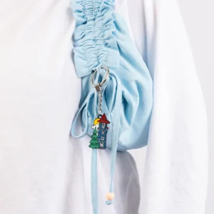 A person in a white top carries a light blue ruched bag with the Moomin Keyring - Moomin House attached, featuring Moomin characters, a colourful house, and playful charms like a green tree and white bird. Gate21.ee