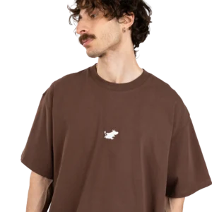A man with curly brown hair and a moustache wears the Moomin Heavy Ts - Chocolate, a loose-fitting brown T-shirt featuring a small white Moomin graphic on the chest, standing against a plain white background. Gate21.ee