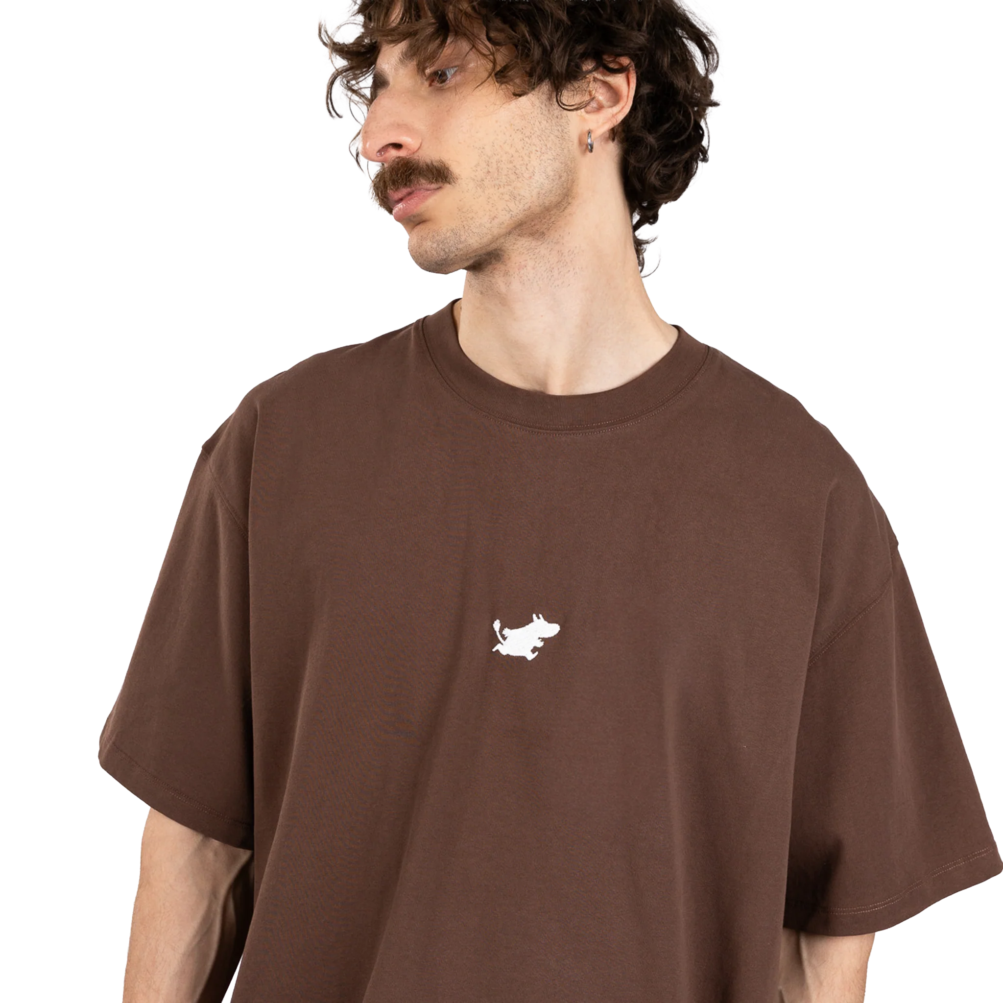 A man with curly brown hair and a moustache wears the Moomin Heavy Ts - Chocolate, a loose-fitting brown T-shirt featuring a small white Moomin graphic on the chest, standing against a plain white background. Gate21.ee