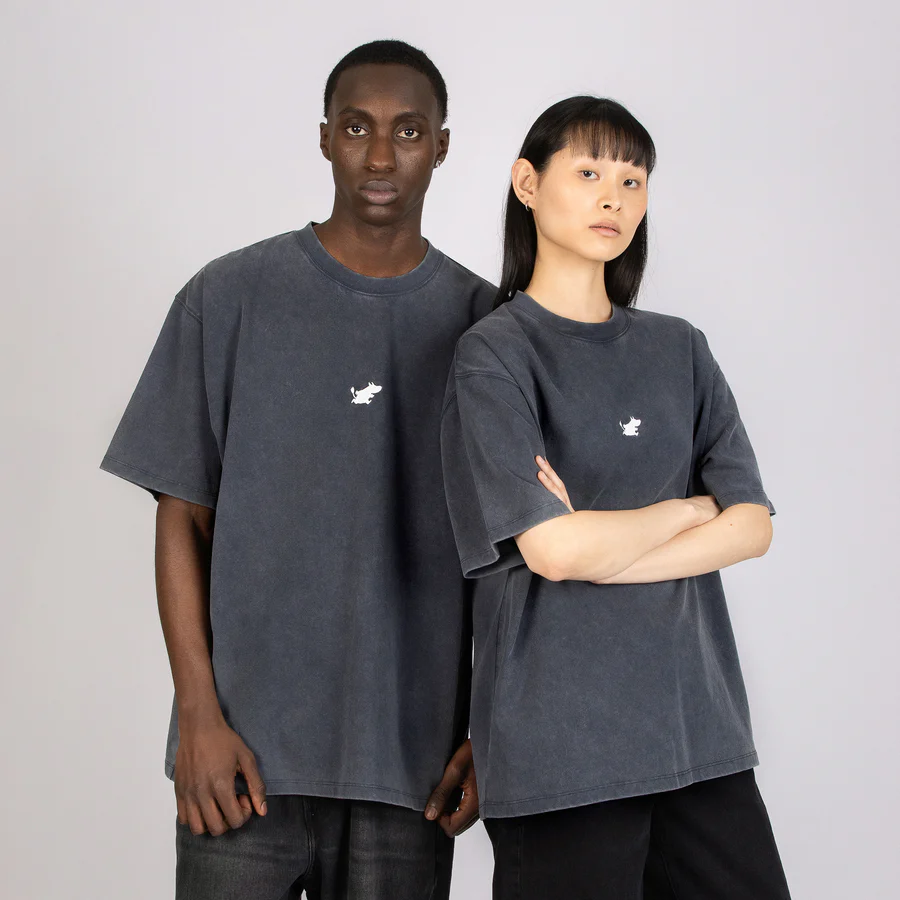 Two people stand side by side against a neutral background, both wearing loose-fitting Moomin Ts in Stone Wash Blue with a small white chest graphic. The person on the right has their arms folded. Gate21.ee