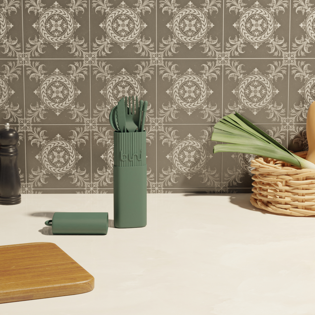The Bini Cutlery Kit - Green stands on a kitchen worktop near its matching case, beside a wooden chopping board, a vegetable basket, a pepper mill, and a splashback with ornate grey tile patterns. Gate21.ee
