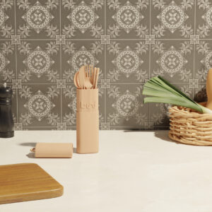 The Bini Cutlery Kit - Nude, featuring wooden utensils in a beige holder, rests on a white worktop before grey and white patterned tiles. A wicker basket with vegetables and a pepper mill sit nearby. Gate21.ee