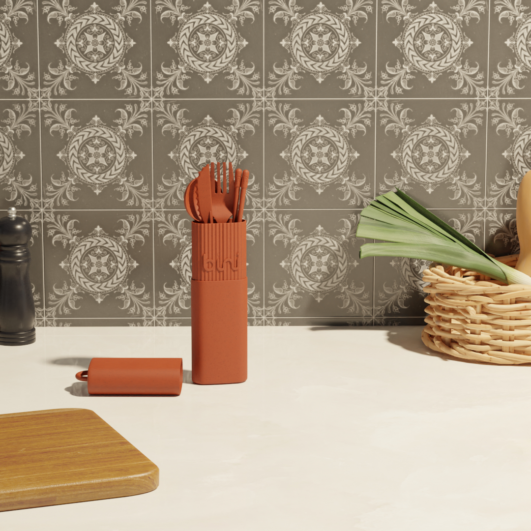 The Bini Cutlery Kit - Terracotta sits on a light kitchen worktop with a basket, leek, pepper mill, small brown container, wooden board, and patterned grey tiles in the background. Gate21.ee
