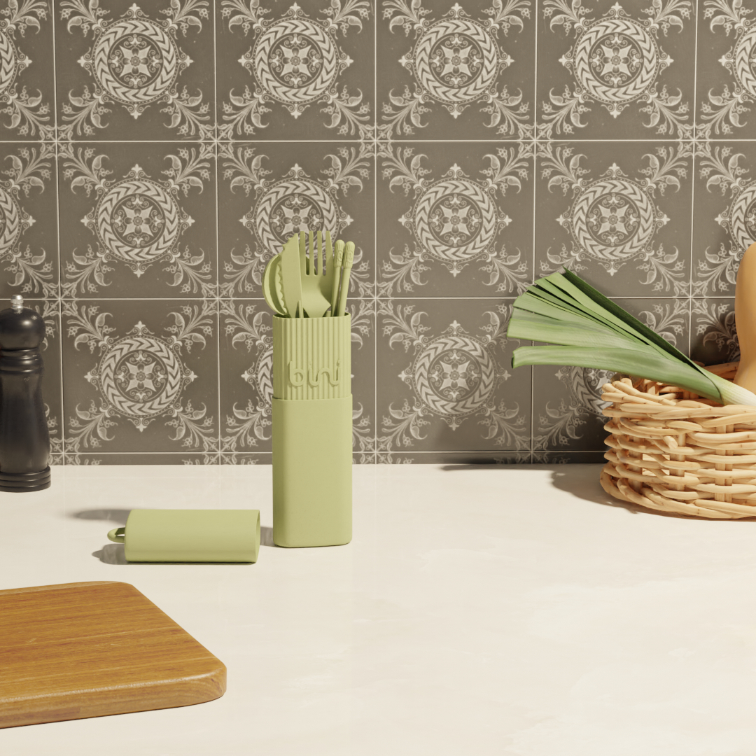 The Bini Cutlery Kit - Olive stands upright in its matching case on a kitchen worktop, set against patterned grey tiles. Nearby are a wicker vegetable basket, a salt or pepper grinder, and a wooden board. Gate21.ee