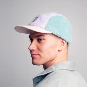 A person with short blond hair, wearing the Moomin Fleece Cap - Multicolour featuring keyrings and a light collared shirt, faces left against a plain grey background. Gate21.ee