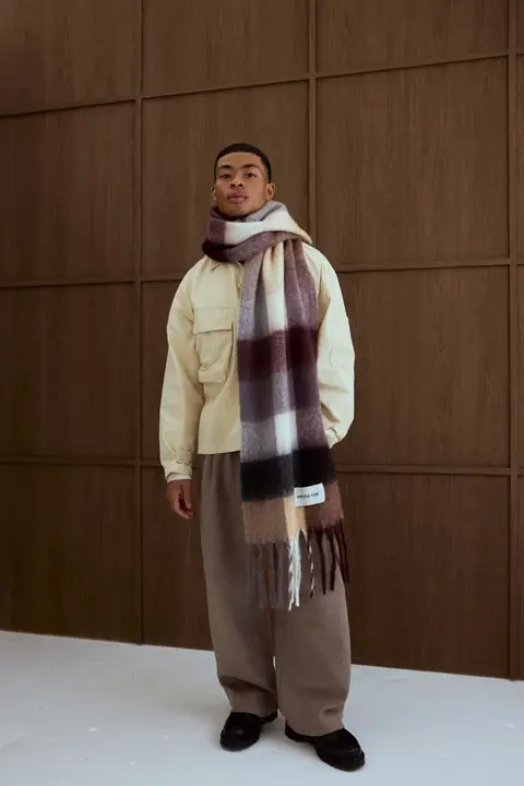 A person stands indoors by a wood-panelled wall, wearing a light beige shirt, brown wide-leg trousers, black shoes, and The Reykjavik Scarf in Purple/Brown draped around their neck. Gate21.ee