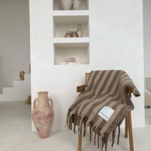 A wooden chair draped with the Wool Stripe Throw - Burnt Umber sits before built-in white shelves displaying ceramic vases and containers, with a large beige ceramic vase placed on the floor nearby. Gate21.ee